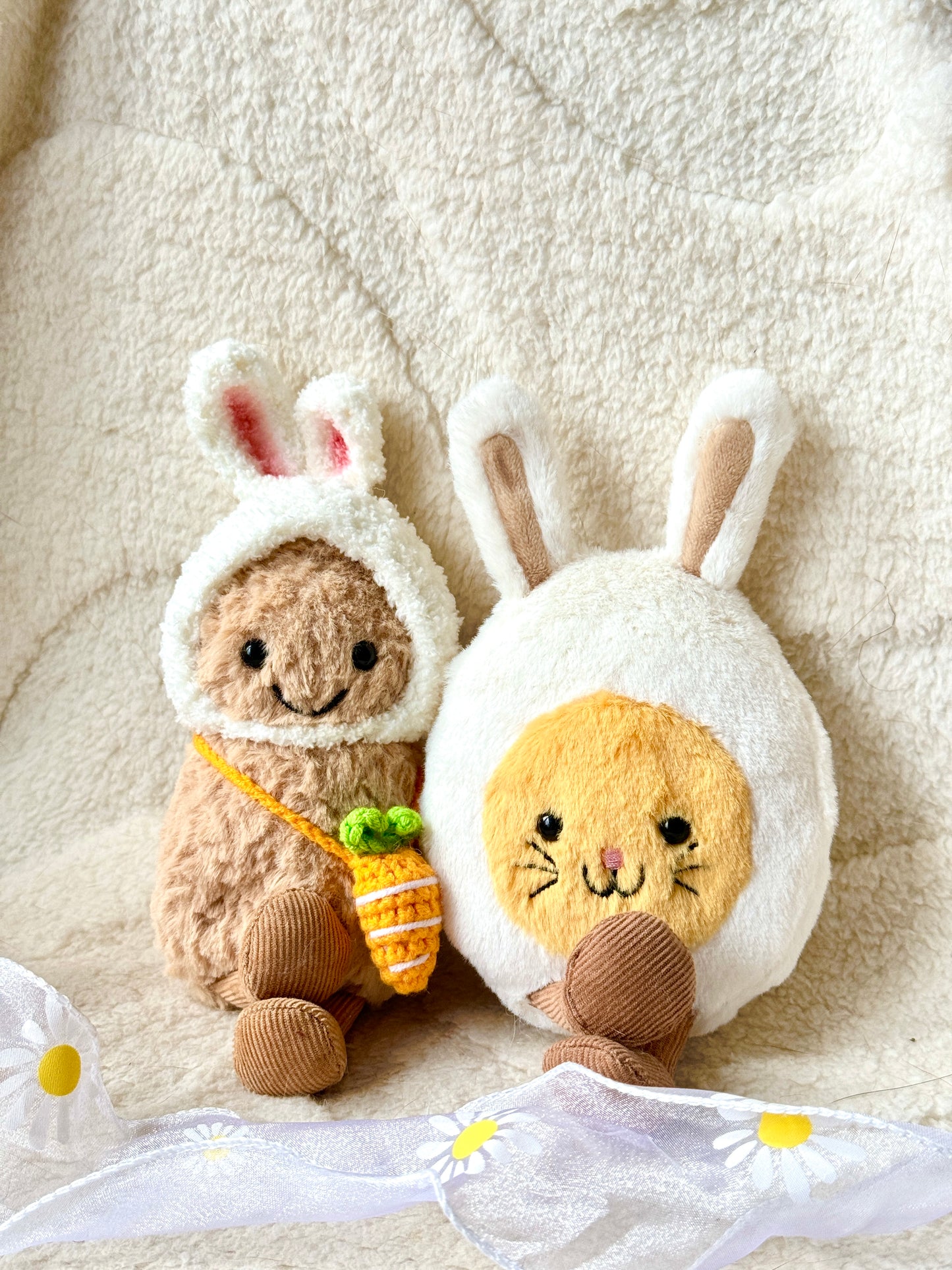 A Jellycat Little Peanut plush wearing a handmade fluffy white bunny hat with pink-lined ears and a small crochet carrot bag slung across its body. The plush sits next to a Jellycat bunny egg plush, both placed on a soft, neutral-toned fabric background with delicate white daisy ribbon accents.