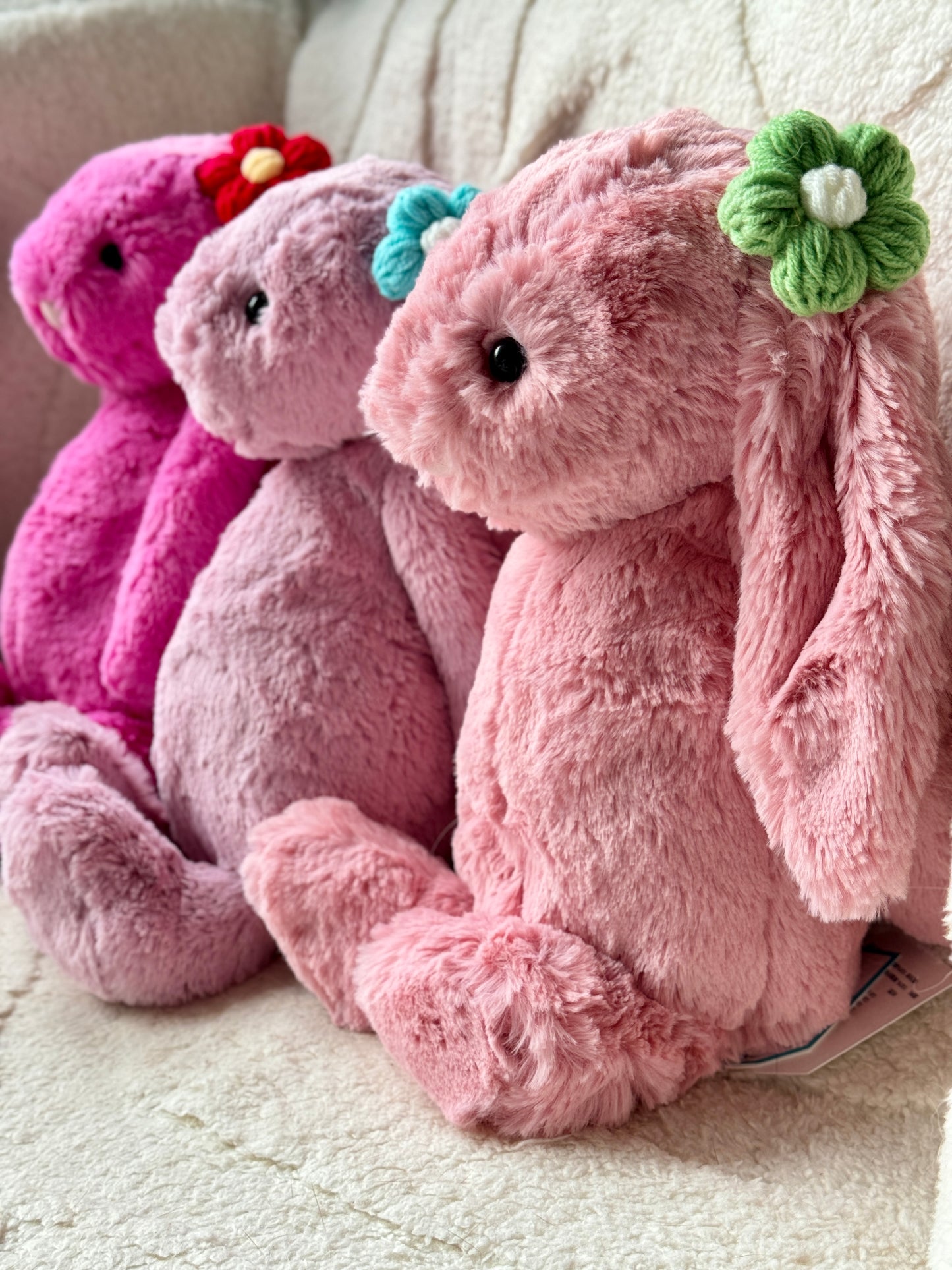 Three Jellycat Bashful Bunnies in shades of pink, sitting in a row on a soft cream-colored surface. Each bunny wears a handmade crochet flower hair accessory in different colorsโgreen, blue, and red.