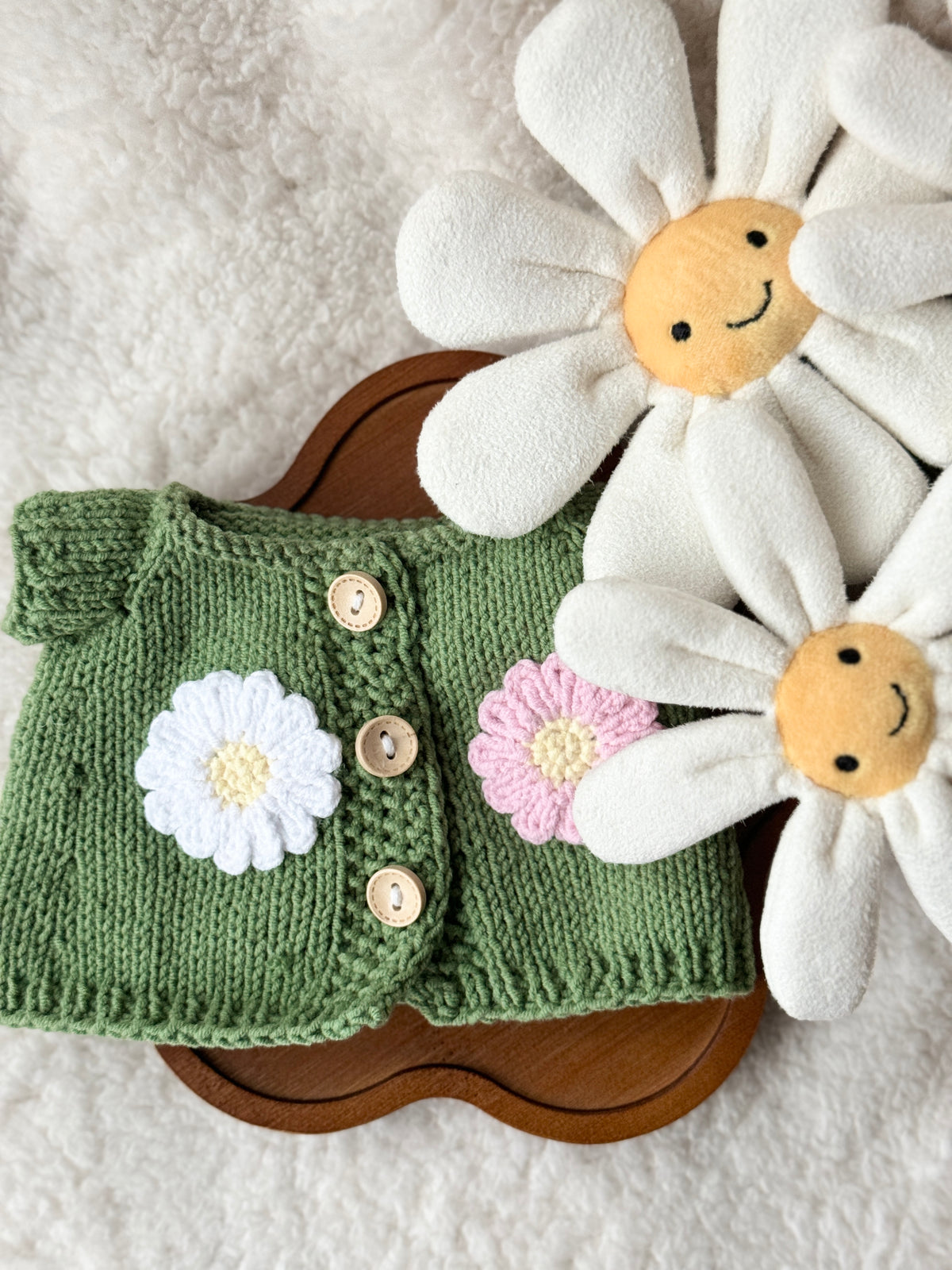 Flat lay of handmade sage green knit cardigan for Jellycat Bashful Bunny with white and pink crochet daisy appliqués and natural wooden buttons on wooden tray