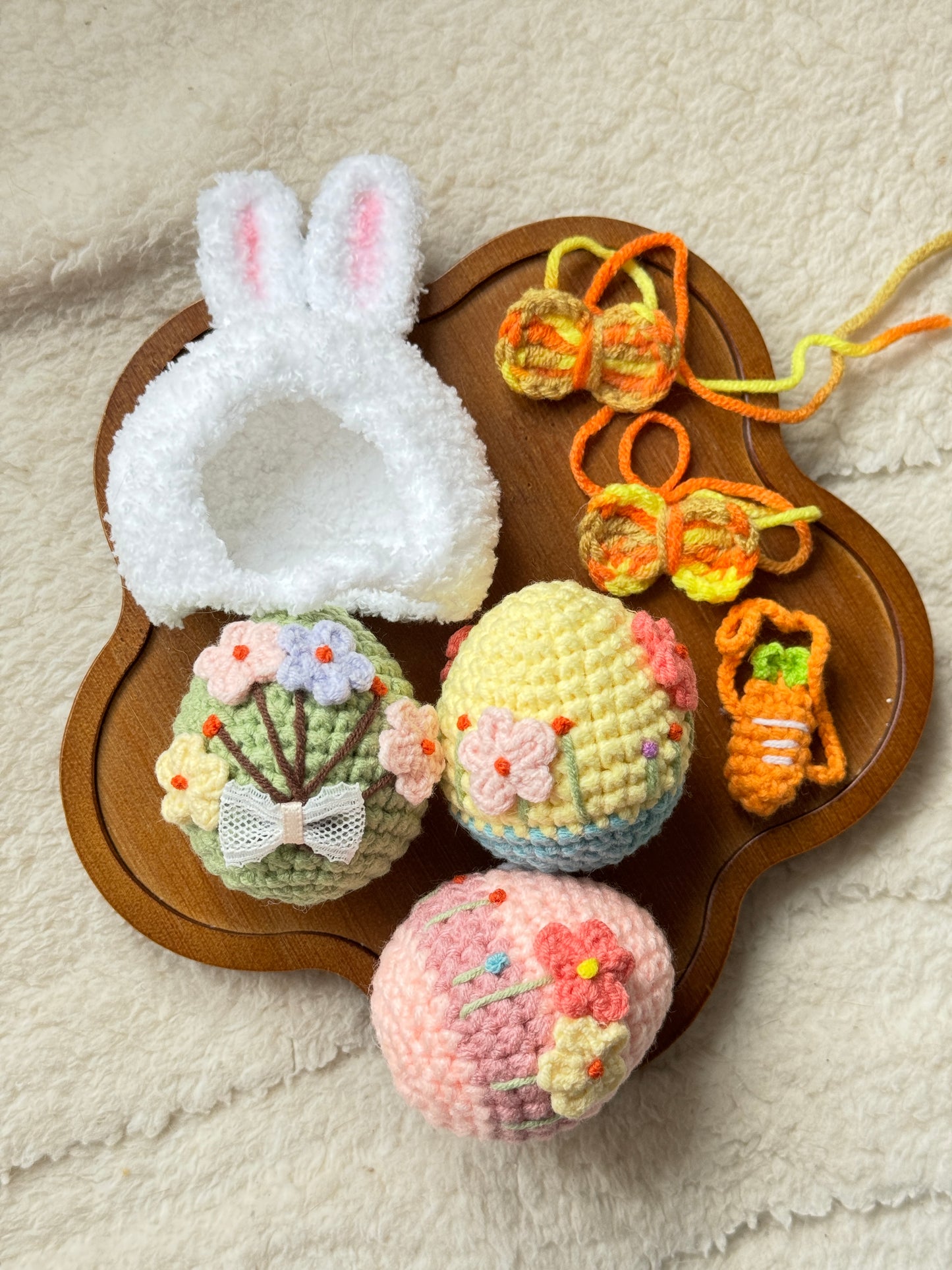 Flat lay of handmade Easter-themed plush accessories including a white bunny hood, orange crochet carrot necklace, two butterfly hair clips, and three embroidered crochet Easter eggs with floral designs, arranged on a wooden tray.