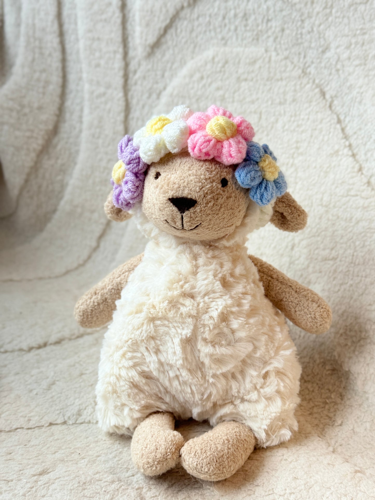 A soft, plush Jellycat sheep wearing a handmade crochet floral headband in pastel colorsโwhite, pink, blue, and lavender flowers with yellow centers. The headband adds a dreamy, fairycore touch to the plush.