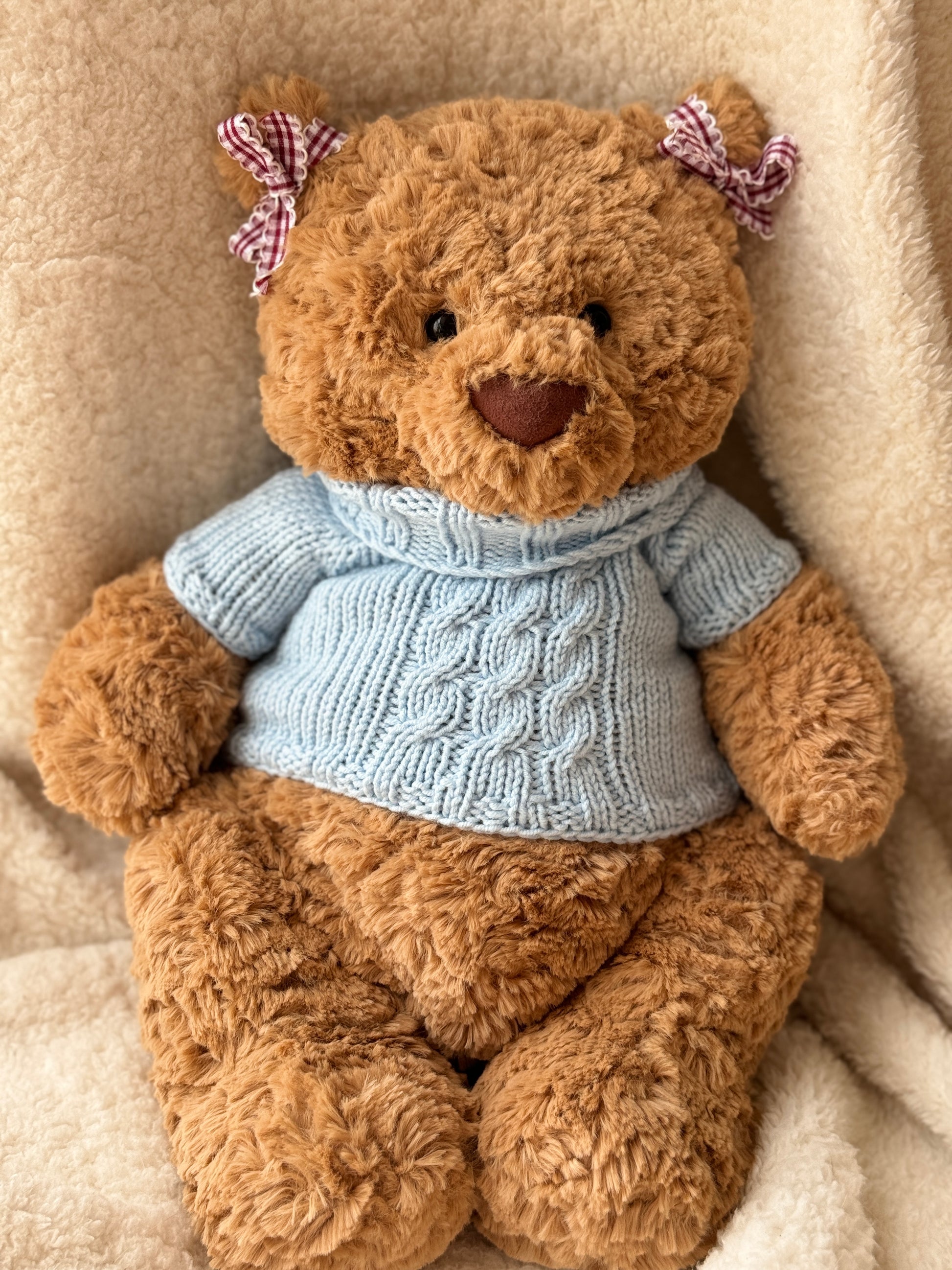 Jellycat Bartholomew Bear wearing a handmade blue cable knit sweater, styled on a soft cream blanket with a cozy plush look.