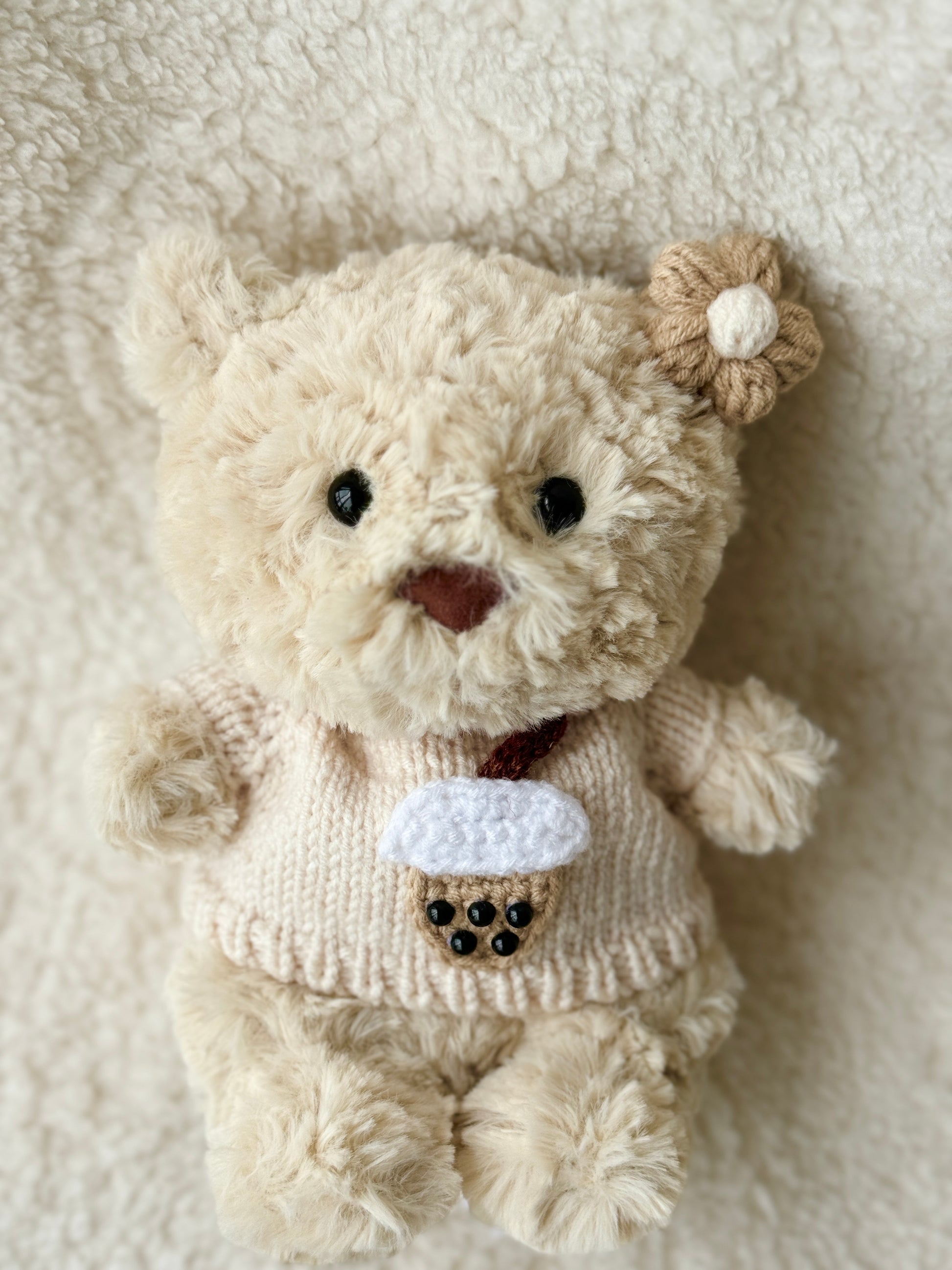 Top-down view of Jellycat Bartholomew Junior in handmade boba tea sweater and crochet flower clip, laid on fluffy background