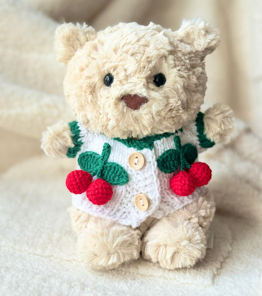 Jellycat Bart Junior wearing a handmade white cherry cardigan with green trim, red crochet cherries, and button details on a soft cream blanket.
