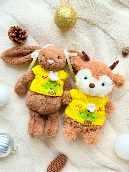 Jellycat Little Bunny and Little Reindeer wearing matching yellow Christmas tree jumpers on a cream blanket with baubles and pinecones.