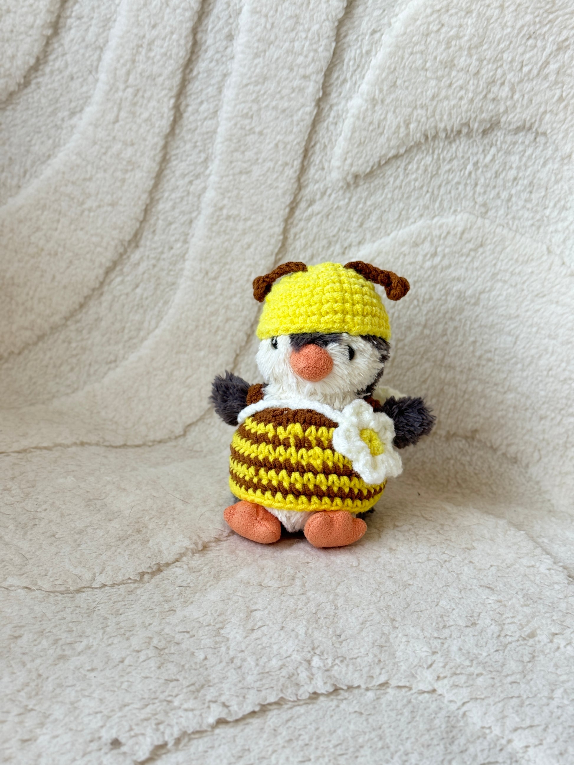 Jellycat Penguin plush dressed in a handmade crochet bee outfit, featuring a yellow and brown striped dress, a yellow hat with brown antennae, and a white flower crossbody bag. Posed solo against a cream textured fabric background.

