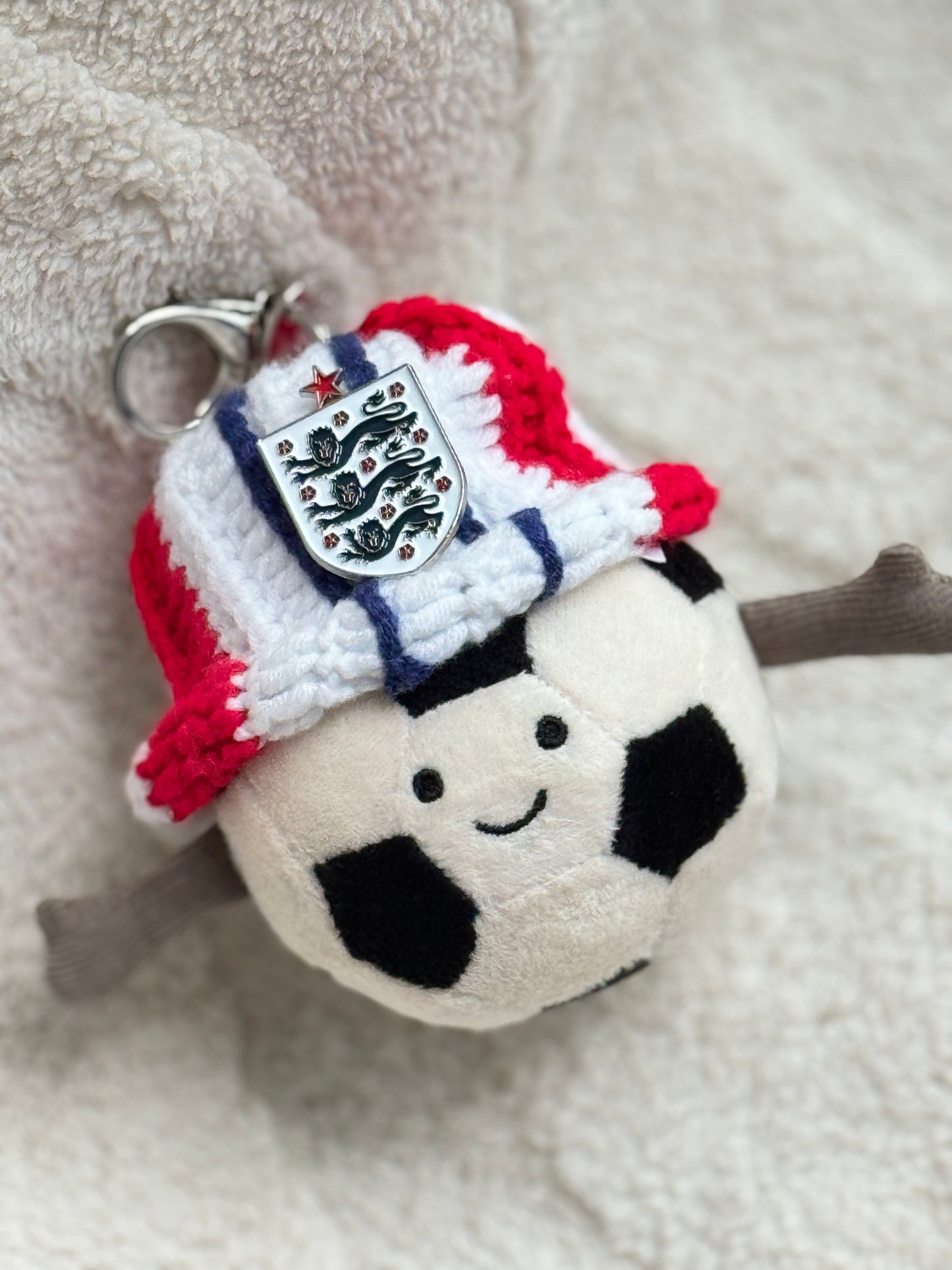 Jellycat Amuseable Sports Football plush wearing a handmade England supporter crochet bucket hat in red, white and blue with an England Three Lions badge attached. Cozy football fan aesthetic inspired by World Cup and England football culture.