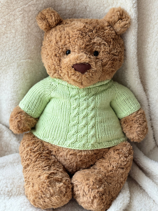 Jellycat Bartholomew Bear wearing a handmade green cable knit sweater, styled on a soft cream blanket with a cozy plush look.