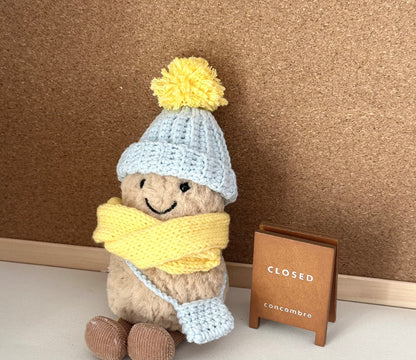 A Jellycat Amuseable plush dressed in a handmade crochet winter outfit. The light blue pom-pom beanie and matching crossbody bag pair perfectly with a vibrant yellow scarf. 