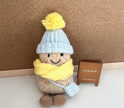 A Jellycat Amuseable plush dressed in a handmade crochet winter outfit. The light blue pom-pom beanie and matching crossbody bag pair perfectly with a vibrant yellow scarf. 