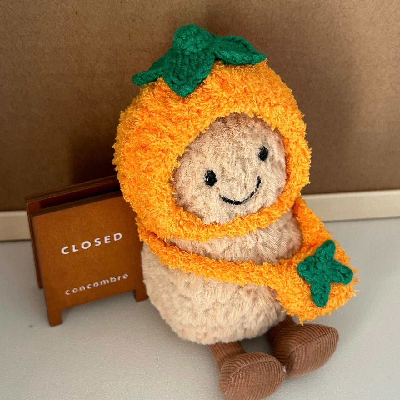 A Jellycat Amuseables plush dressed in a handmade crochet orange outfit, featuring a vibrant orange hood with a green leafy top and a matching bag.