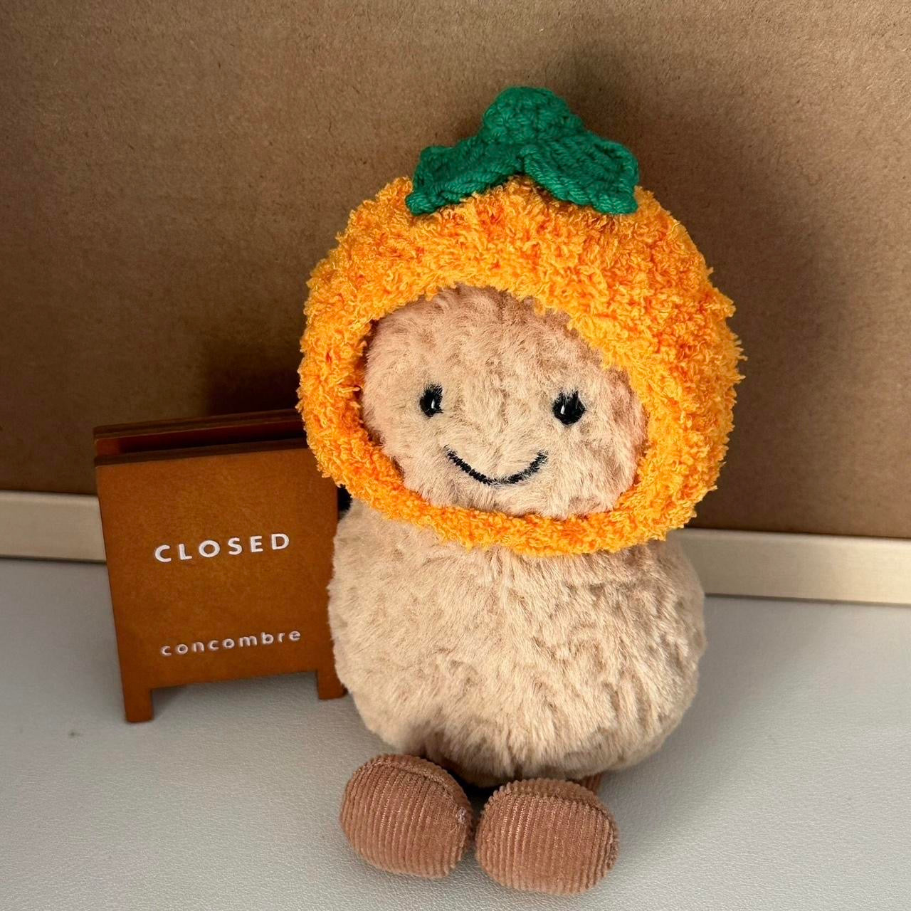 A Jellycat Amuseables plush dressed in a handmade crochet orange outfit, featuring a vibrant orange hood with a green leafy top and a matching bag.