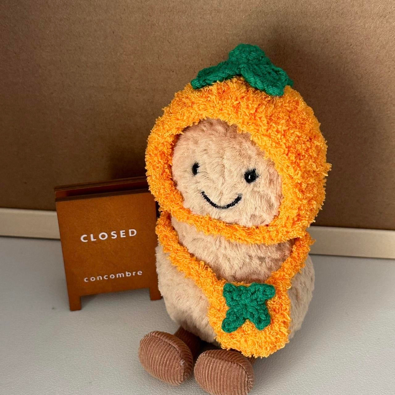 A Jellycat Amuseables plush dressed in a handmade crochet orange outfit, featuring a vibrant orange hood with a green leafy top and a matching bag.