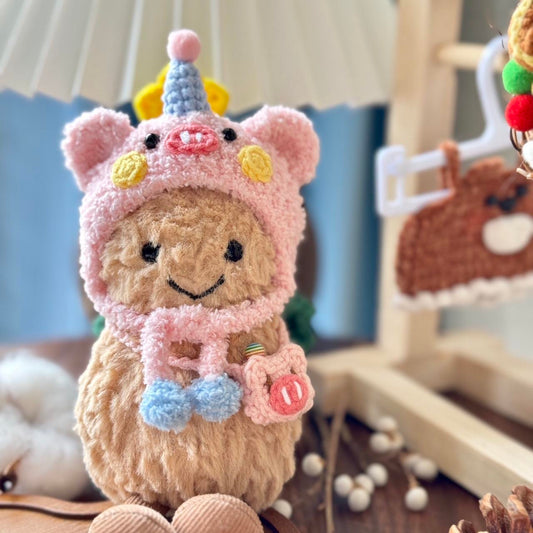 A Jellycat Amuseables plush dressed in a handmade crochet piggy party outfit, featuring a pink hood with pig ears, a snout, rosy cheeks, a pastel blue and yellow party hat, and a matching small pink pig-shaped bag. The plush sits on a wooden table with a cozy background.