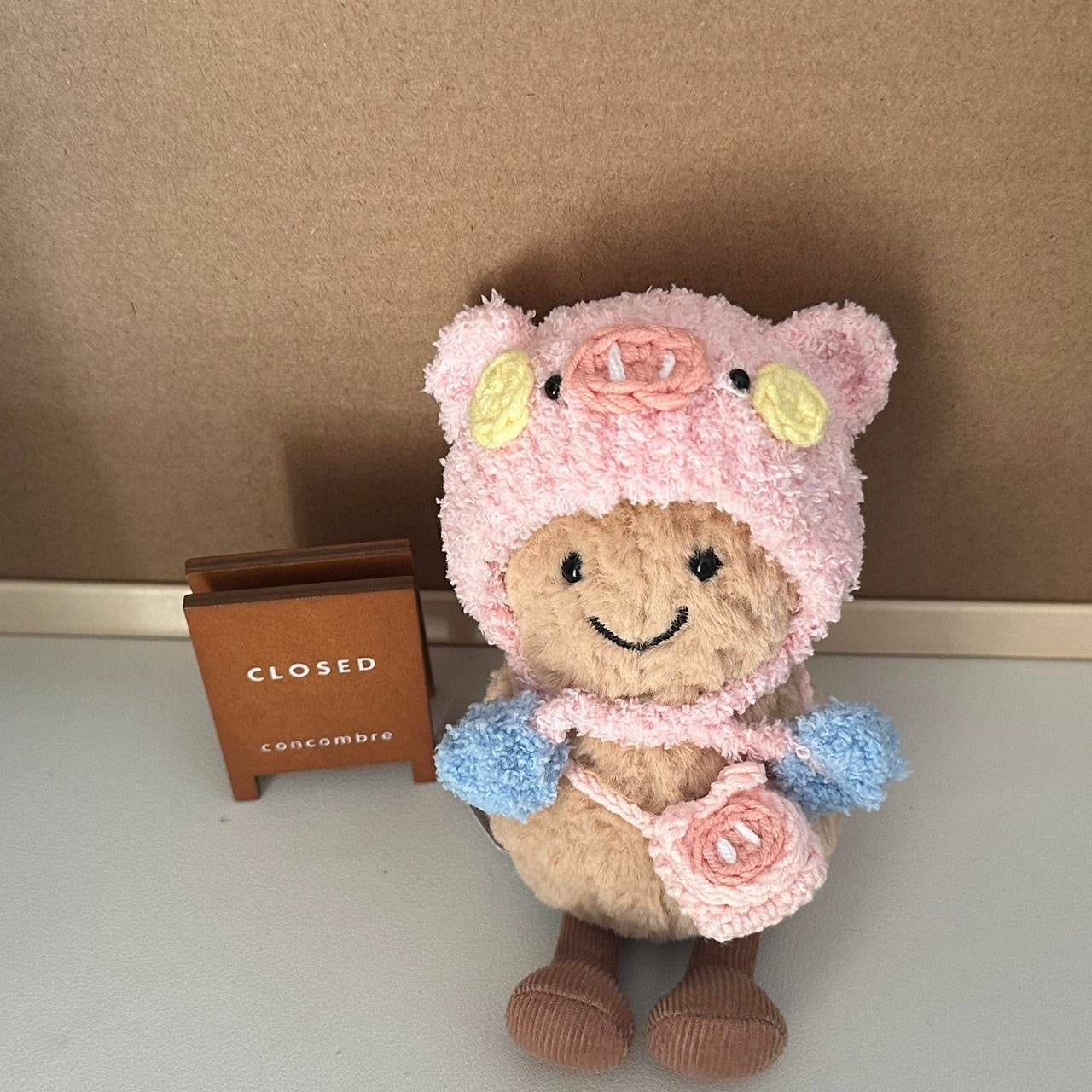 A Jellycat Amuseables plush dressed in a handmade crochet piggy party outfit, featuring a pink hood with pig ears, a snout, rosy cheeks, a pastel blue and yellow party hat, and a matching small pink pig-shaped bag. The plush sits on a wooden table with a cozy background.