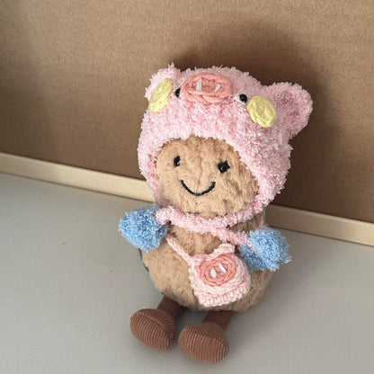 A Jellycat Amuseables plush dressed in a handmade crochet piggy party outfit, featuring a pink hood with pig ears, a snout, rosy cheeks, a pastel blue and yellow party hat, and a matching small pink pig-shaped bag. The plush sits on a wooden table with a cozy background.