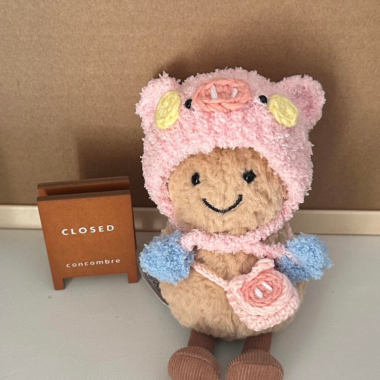 A Jellycat Amuseables plush dressed in a handmade crochet piggy party outfit, featuring a pink hood with pig ears, a snout, rosy cheeks, a pastel blue and yellow party hat, and a matching small pink pig-shaped bag. The plush sits on a wooden table with a cozy background.