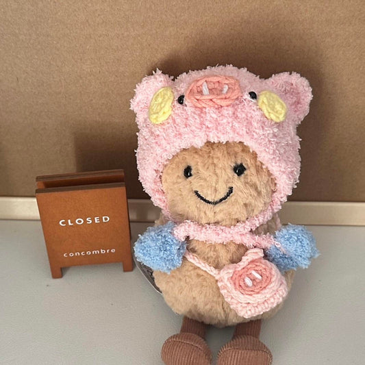 A Jellycat Amuseables plush dressed in a handmade crochet piggy party outfit, featuring a pink hood with pig ears, a snout, rosy cheeks, a pastel blue and yellow party hat, and a matching small pink pig-shaped bag. The plush sits on a wooden table with a cozy background.