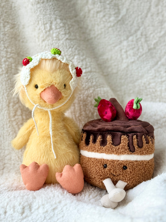 Jellycat Dorit Duck wearing a handmade crochet strawberry triangle bonnet with tiny red strawberry details, styled beside a plush chocolate strawberry cake on a soft cream blanket.