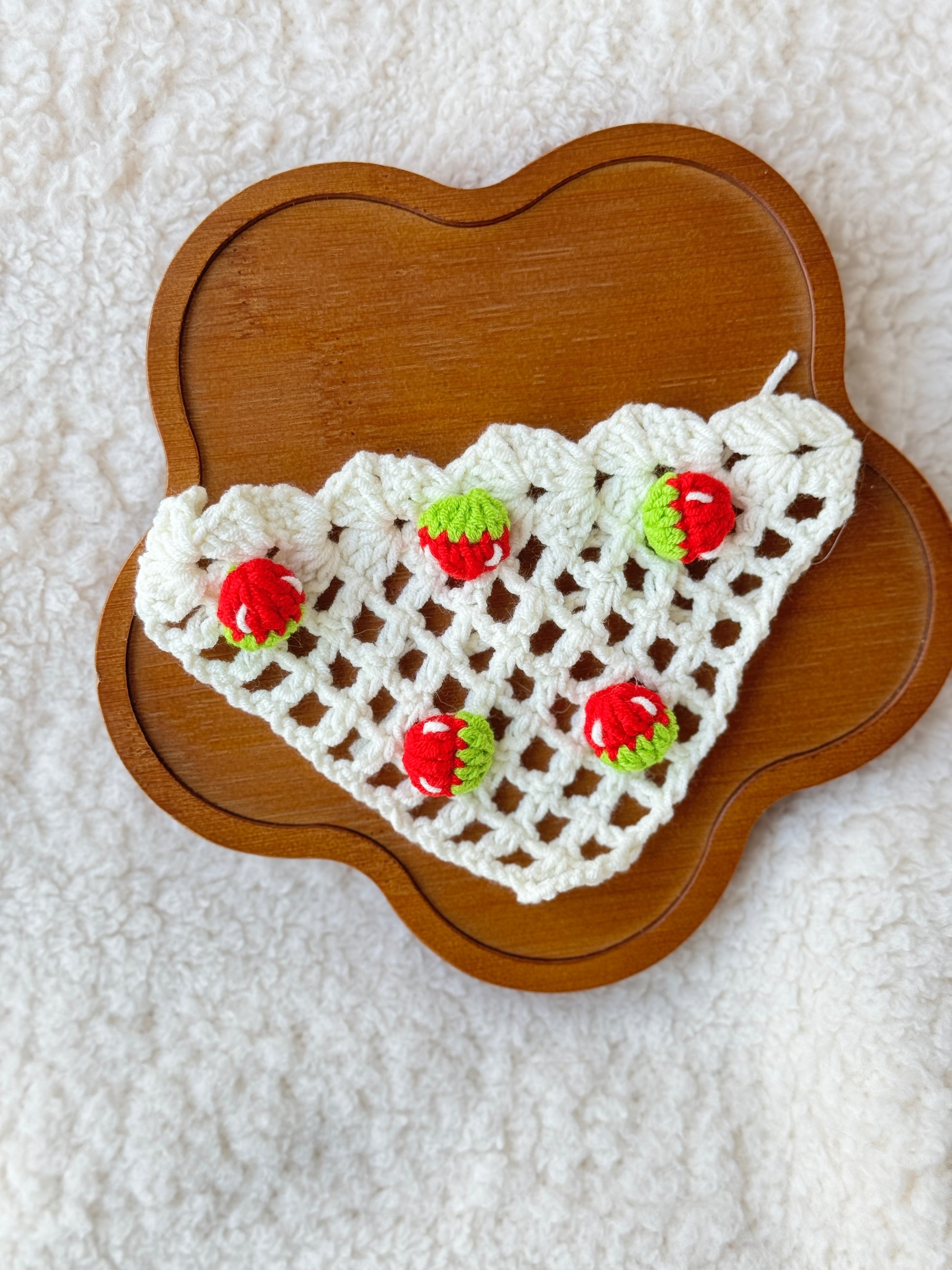 Flat lay of handmade crochet strawberry triangle bonnet on a wooden tray, showing the cream crochet mesh pattern and tiny strawberry details for Jellycat Dorit Duck.