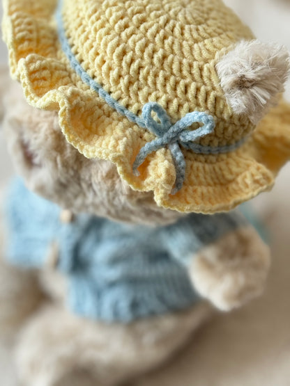 Detail shot of a handmade sunny yellow bonnet for Jellycat Bart Junior, showing the crochet texture, ruffled brim, and pale blue bow.