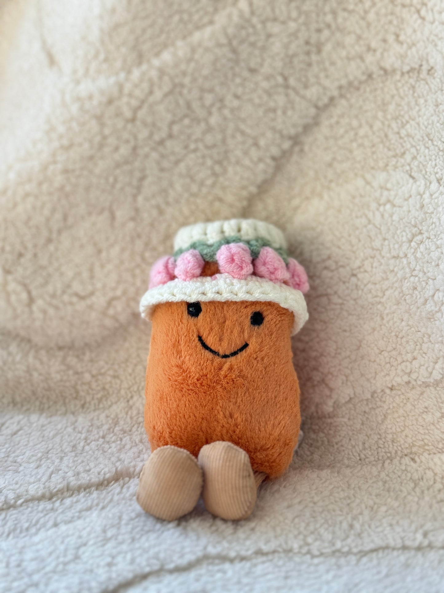 Jellycat Bean plush wearing a handmade crochet tulip flower hat with pink petals and sage green leaf details in a soft cozy cream background.