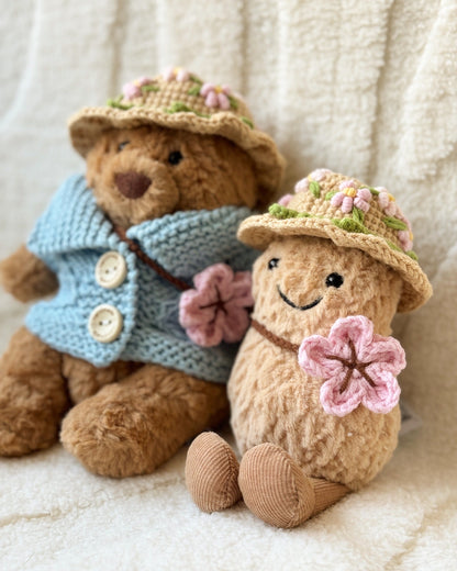 Two Jellycat plush toys wearing handmade crochet outfits. The Bartholomew Bear is dressed in a soft blue cardigan with buttons and a beige floral crochet hat, while the Amuseable Peanut wears a matching floral crochet hat and holds a pink crochet flower. Both outfits feature delicate spring blossom designs, highlighting the craftsmanship and charm of the handmade accessories