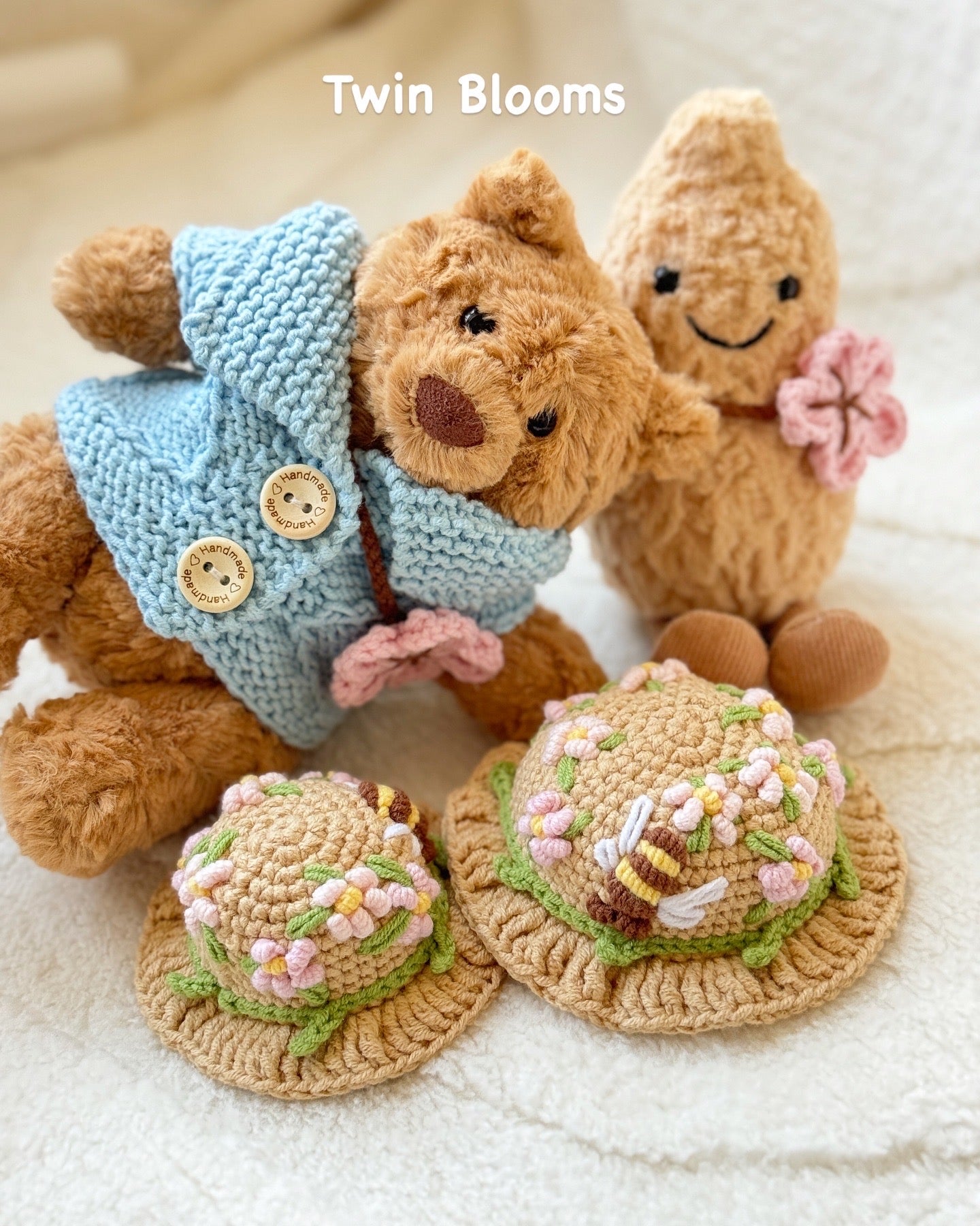 Two Jellycat plush toys wearing handmade crochet outfits. The Bartholomew Bear is dressed in a soft blue cardigan with buttons and a beige floral crochet hat, while the Amuseable Peanut wears a matching floral crochet hat and holds a pink crochet flower. Both outfits feature delicate spring blossom designs, highlighting the craftsmanship and charm of the handmade accessories
