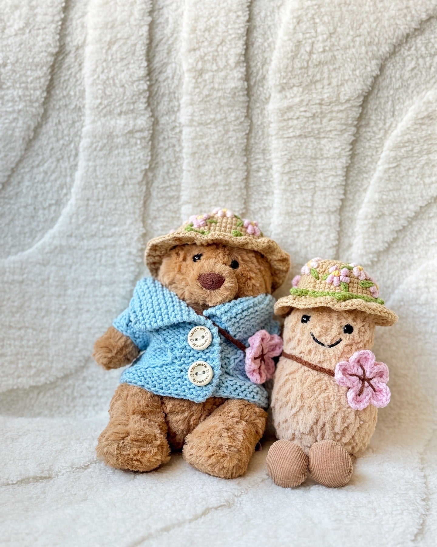 Two Jellycat plush toys wearing handmade crochet outfits. The Bartholomew Bear is dressed in a soft blue cardigan with buttons and a beige floral crochet hat, while the Amuseable Peanut wears a matching floral crochet hat and holds a pink crochet flower. Both outfits feature delicate spring blossom designs, highlighting the craftsmanship and charm of the handmade accessories