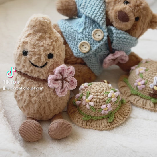 Two Jellycat plush toys wearing handmade crochet outfits. The Bartholomew Bear is dressed in a soft blue cardigan with buttons and a beige floral crochet hat, while the Amuseable Peanut wears a matching floral crochet hat and holds a pink crochet flower. Both outfits feature delicate spring blossom designs, highlighting the craftsmanship and charm of the handmade accessories