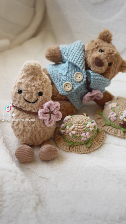 Two Jellycat plush toys wearing handmade crochet outfits. The Bartholomew Bear is dressed in a soft blue cardigan with buttons and a beige floral crochet hat, while the Amuseable Peanut wears a matching floral crochet hat and holds a pink crochet flower. Both outfits feature delicate spring blossom designs, highlighting the craftsmanship and charm of the handmade accessories